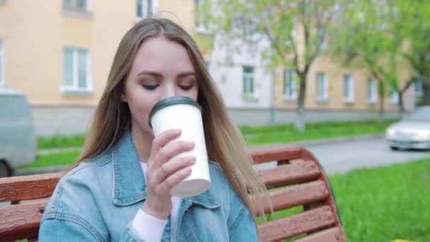 Nápoje a lidé koncept - šťastný mladá žena nebo dospívající dívka pití kávy z papírového kelímku sedí na městské ulici lavičce — Stock video