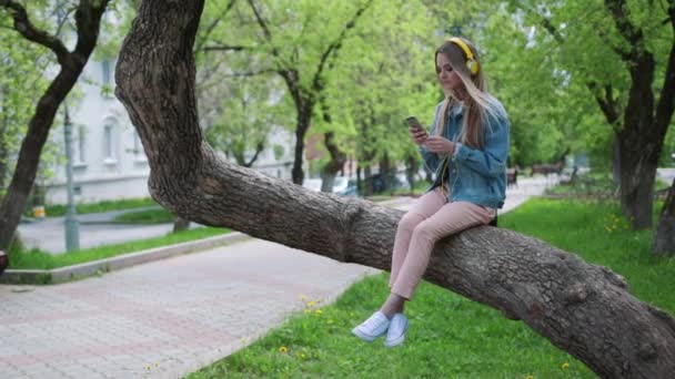 Mujer sentada en un árbol en el parque y escuchando música . — Vídeo de stock