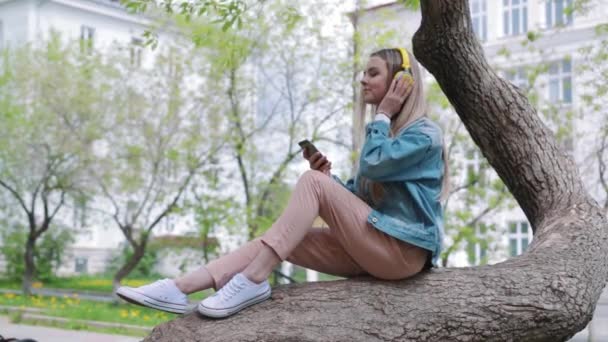 Mujer sentada en un árbol en el parque y escuchando música . — Vídeo de stock