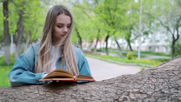 Krásná vážná dívka v džínové bundy a brýle čte knihu, proti letní zelený park — Stock video