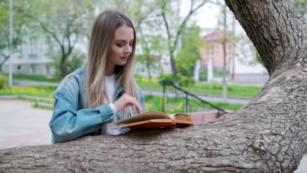 Beautiful serious girl in jeans jacket and glasses reads book, against summer green park — Stock Video