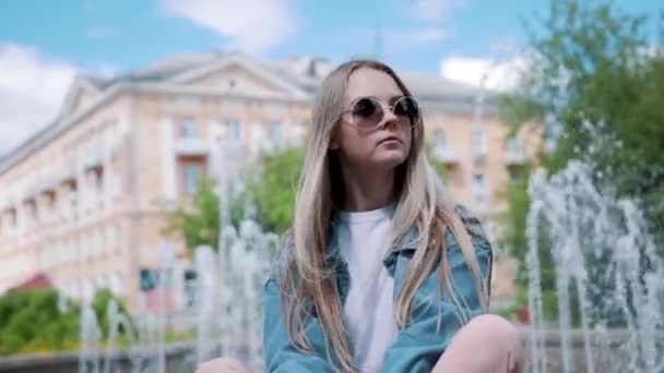 Hermosa joven sentada en un parapeto en el fondo de la fuente, en una chaqueta de mezclilla — Vídeos de Stock