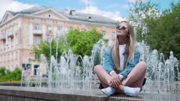 Bella ragazza seduta su un parapetto sullo sfondo della fontana, in giacca di jeans — Video Stock