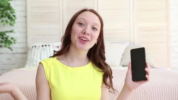 Happy and cool blogger is showing the phone to the vlogging camera. She likes her new present. Young woman is showing the big thumb up. She can recommend this phone to her subcribers. — Stock Video