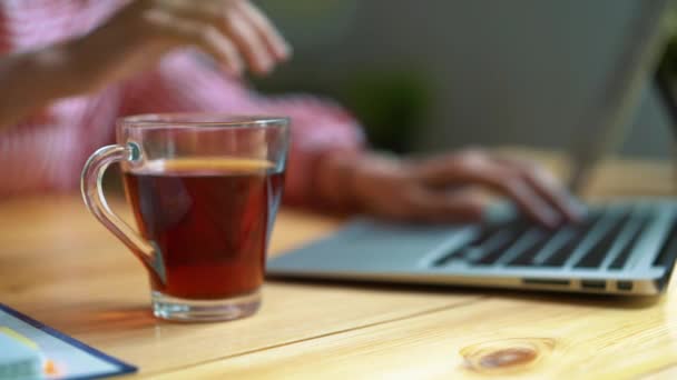 Een meisje drinkt thee tijdens het werken, zitten aan een tafel, een transparante mok thee. — Stockvideo