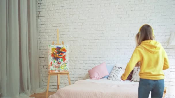 Linda chica se divierte bailando en la cama en casa escuchando música con auriculares saltando y levantando las manos. Estilo de vida moderno y concepto de jóvenes felices . — Vídeos de Stock