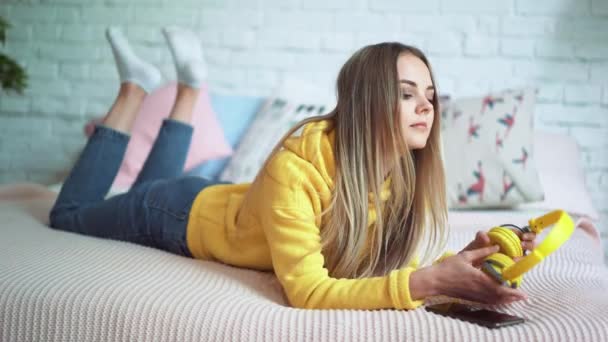 Mooi meisje luisteren naar muziek op de hoofdtelefoon op het bed en ontspannen met behulp van de telefoon — Stockvideo