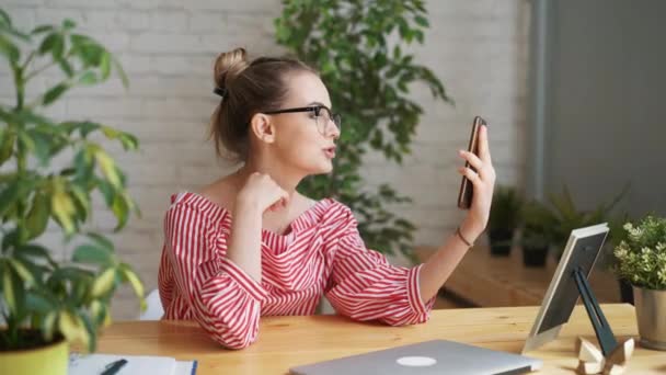 Šťastná žena vysokoškolský student s video volání přes mobil, sedí s notebookem. ženské na volné noze pomocí wifi připojení pro vzdálenou práci a blogování. — Stock video