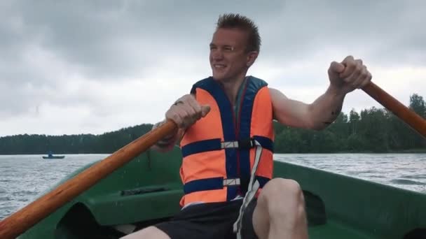 Mladý muž v loďce veslování dřevěná vesla na vodě. Plovací vesta na těle muže. — Stock video