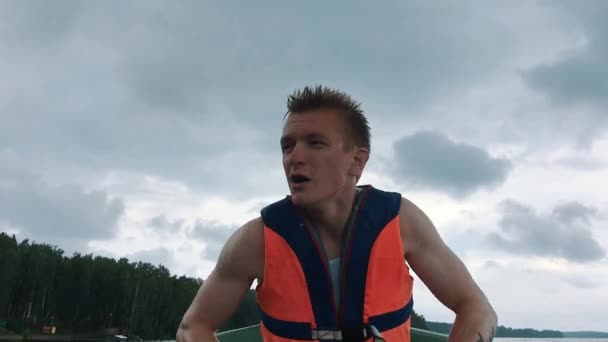 Een jonge man in een boot roeien houten roeiriemen op het water. Zwemvest op het lichaam van een man. — Stockvideo