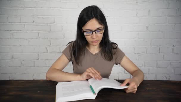 Une fille assise à une table blanche avec des lunettes lisant un livre, un journal intime. Mauvaise vue . — Video