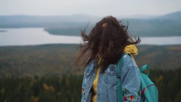 Приваблива жінка стоїть на вершині гори з довгим волоссям, що дме на вітрі посміхаючись і намагаючись тримати волосся від її обличчя рукою — стокове відео