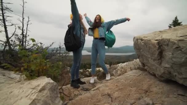 Dos jóvenes mujeres felices bailando y levantando brazos en la hermosa cima de la montaña sobre el cielo del atardecer . — Vídeo de stock