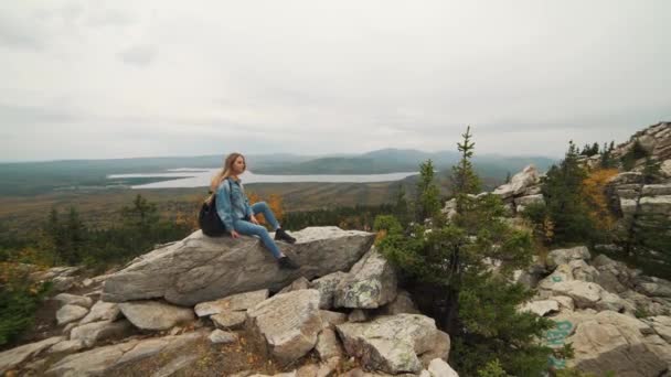 Una joven sentada en la cima de la montaña mirando un hermoso paisaje pensando en la vida. Una chica de vacaciones . — Vídeos de Stock