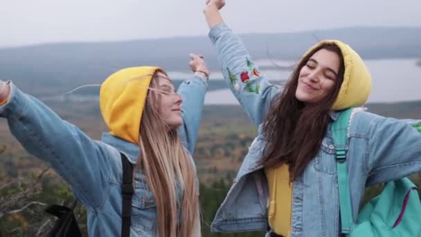 Zwei junge glückliche Frauen tanzen und heben die Arme auf einem wunderschönen Berggipfel über dem Himmel bei Sonnenuntergang. — Stockvideo