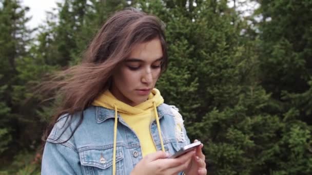 Молодой студент использует сенсорный экран смартфона в то время как в природе. Хипстер пользуется социальными сетями и улыбается, читая мобильные. Красивая женщина в джинсовой куртке в еловом лесу — стоковое видео