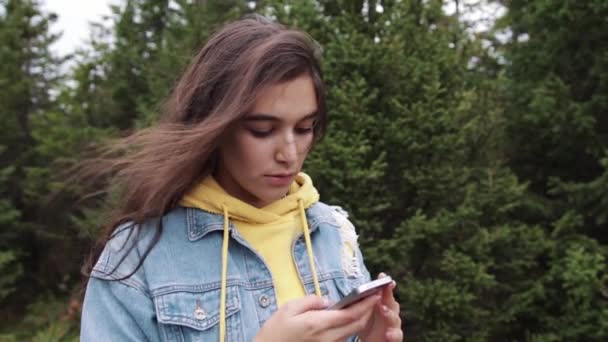 Adolescente joven mixta usando el teléfono inteligente de pantalla táctil mientras está en la naturaleza. Hipster utilizando las redes sociales y sonriendo mientras lee el móvil. Hermosa mujer en chaqueta de mezclilla en el bosque de abeto — Vídeos de Stock