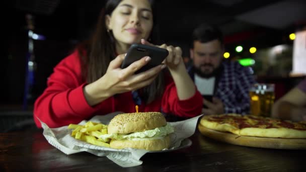 Ένα κορίτσι στην εταιρεία από έναν άνδρα, λαμβάνοντας εικόνες στο smartphone σας Burger σας στο μπαρ. — Αρχείο Βίντεο
