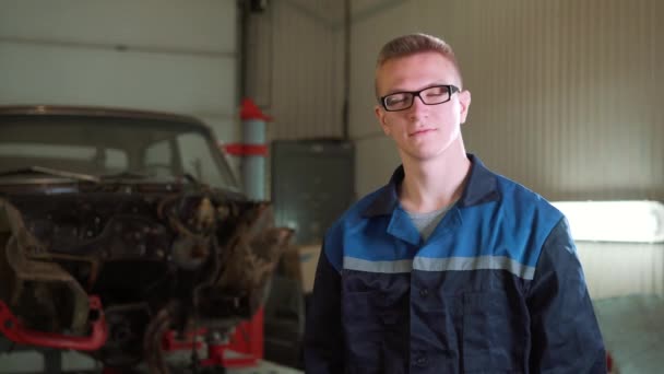 Ritratto di un giovane bellissimo meccanico in officina, sullo sfondo di un'auto rotta. Concetto: riparazione della macchina, diagnosi dei guasti, riparazione specialistica, manutenzione . — Video Stock