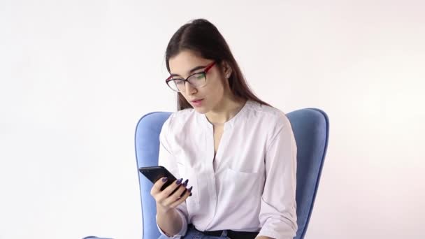 Glimlachend brunette vrouw zitten in een leunstoel en met behulp van een smartphone op witte achtergrond — Stockvideo