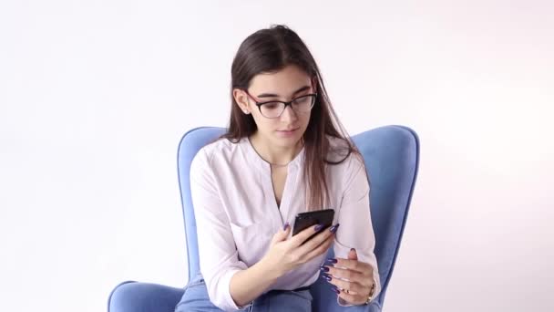 Mujer morena sonriente sentada en un sillón y usando un teléfono inteligente sobre fondo blanco — Vídeos de Stock