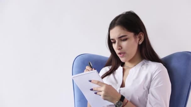 Glimlachend brunette vrouw zitten in een leunstoel en met behulp van een smartphone op witte achtergrond — Stockvideo