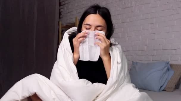 Una joven sentada en casa en la cama, enferma, sonándole la nariz con un pañuelo — Vídeos de Stock