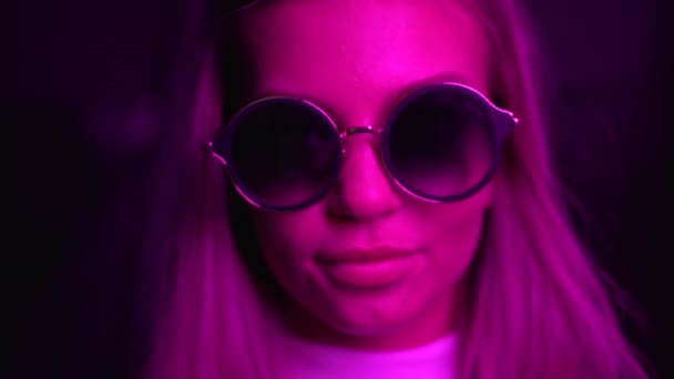 A menina olha para a câmera, tira e põe óculos de sol. Luz de néon — Vídeo de Stock