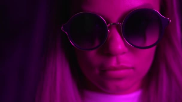 Blickt das Mädchen in die Kamera, zieht die Sonnenbrille auf. Neonlicht — Stockvideo