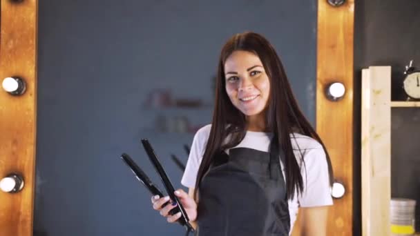 Portret a mosolygó szép nő fodrász, fekete kötényt keres a kamera, miközben professzionális fodrászeszközök szalon háttér — Stock videók
