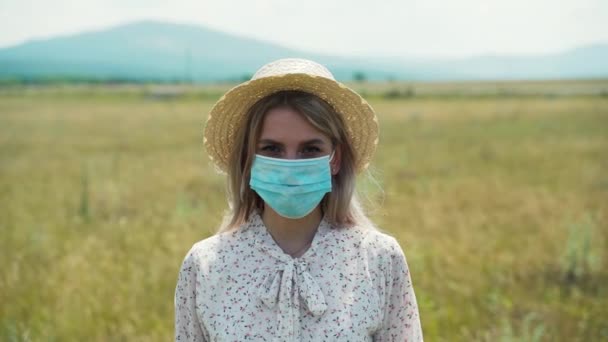 Een meisje staat in een veld, met een medisch masker naar de camera te kijken. — Stockvideo