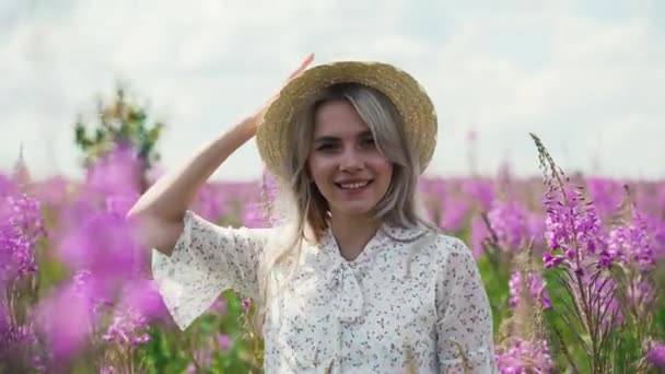 Una chica de pelo rubio con un vestido blanco de verano se para en un campo de té floreciente y se quita el sombrero. . — Vídeos de Stock