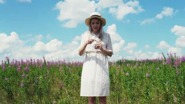 Счастливая милая девушка в белом платье и шляпе медленно срывает листья с ромашки, стоя на горизонте — стоковое видео