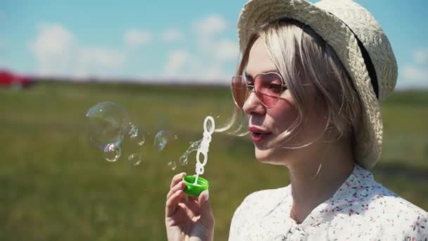 A fair-haired girl in a white summer dress and hat stands in a field of blooming tea and inflates soap bubbles. — Stock Video