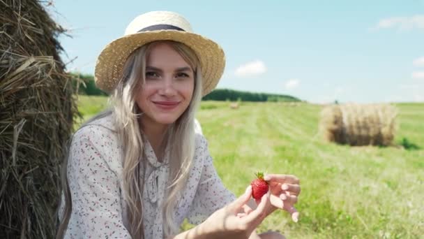 Красивая молодая девушка в белом платье и шляпе сидит в поле и ест клубнику. — стоковое видео