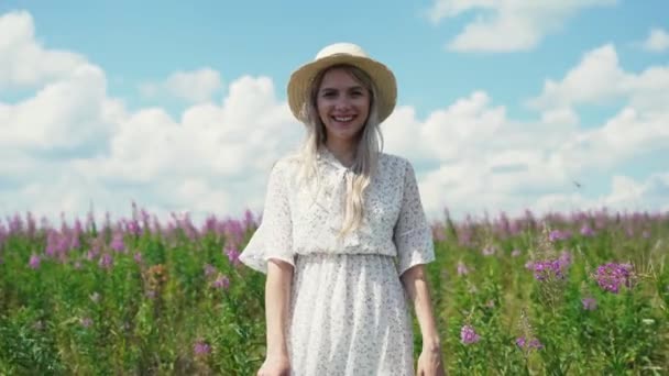 Χαριτωμένο κορίτσι σε ένα λευκό φόρεμα και ένα καπέλο στέκεται ήρεμα σε ένα πεδίο ανθίζοντας τσάι και χαμογελώντας. — Αρχείο Βίντεο