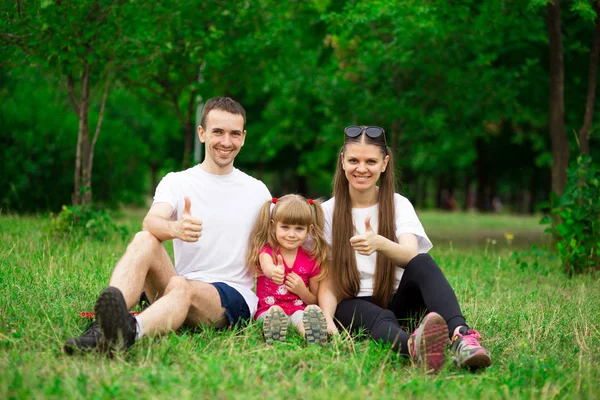 Happy young family of father, mother and little girl sit on green grass in park, enjoying walk in natural environment
