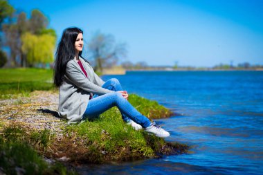 Kot pantolon güzel genç kadın nehir kıyısında güneşli bir günde oturur.