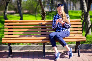 Parktaki bankta otururken cep telefonu kullanan genç bir kadın.