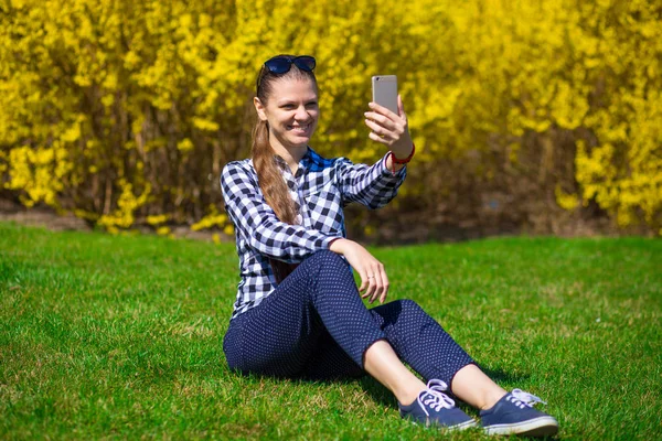 Güzel kız Park'ta rahatlayın ve selfie tree yakınındaki yeşil çimenlerin üzerinde sarı çiçek al. Bahar zamanı. İletişim kavramı