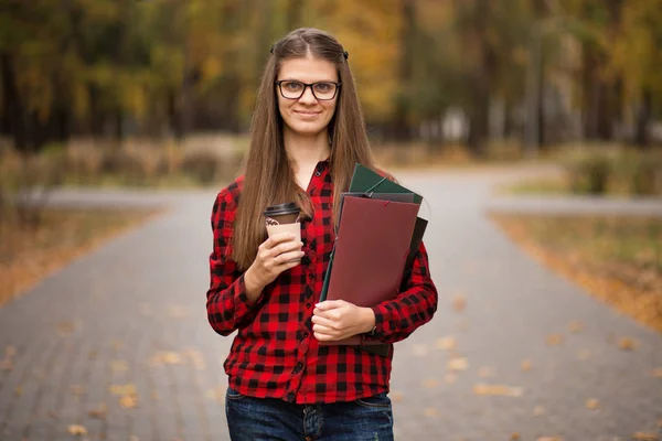 Young student in red checkered shirt with cup of coffee. Portrait of beautiful young woman holding folders in autumn park