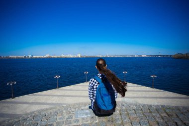 Turist kadının güneşli günde pier deniz kenarında oturan sırt çantası ile arkadan görünüm