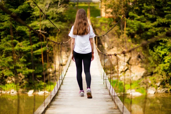Turystyczna kobieta chodzić przez długi, drewniany most wiszący nad rzeką. — Zdjęcie stockowe
