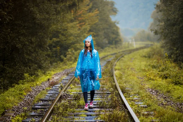 Girl traveller wearing blue jacket and go by forest railway. Autumn and raining season with dark green tones while girl in blue rain jacket walks in forest, fog