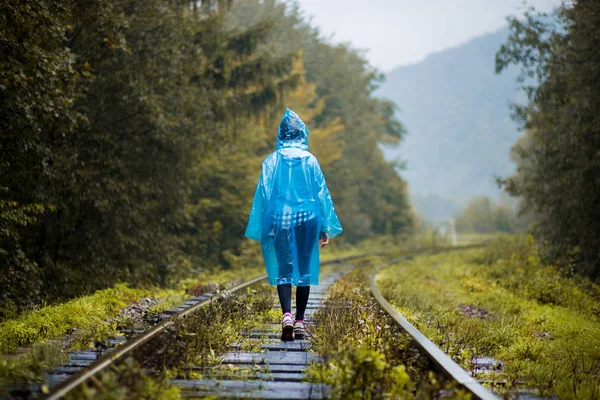 Girl traveller wearing blue jacket and go by forest railway. Autumn and raining season with dark green tones while girl in blue rain jacket walks in forest, fog, rain and clouds — Stock Photo, Image