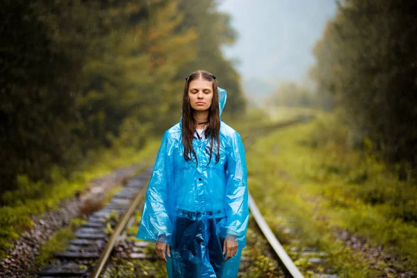 Girl traveller wearing blue jacket and go by forest railway. Autumn and raining season with dark green tones while girl in blue rain jacket walks in forest, fog, rain and clouds