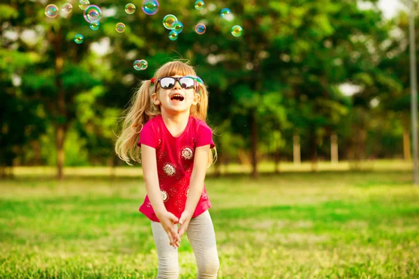 Happy little blond girl blows soap bubbles in summer park — Stock Photo, Image