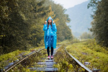 Kız Gezgin giyiyor mavi ceket ve go orman tren tarafından. Mavi yağmur ceket kız ormanda, sis yürür iken koyu yeşil tonları ile sonbahar, yağmur sezonu