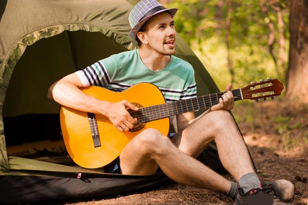 Joven guapo atractivo barbudo modelo hombre en sombrero tocando la guitarra en el bosque . — Foto de Stock