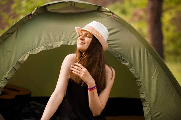 Young slim hiker woman sitting at small tourist tent enjoying beautiful nature forest on bright summer morning.